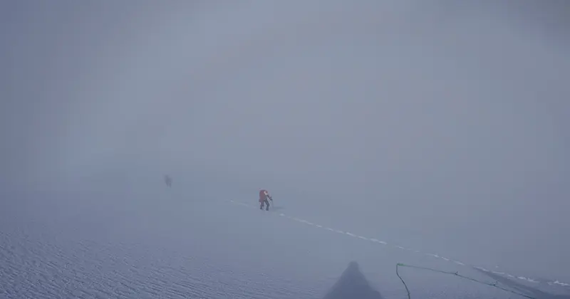 Twee bergbeklimmers lopen door een besneeuwd, mistig landschap en laten voetafdrukken achter. Door de dichte mist is het zicht slecht, en boven hen verschijnt in de mist een vage, regenboogachtige boog.